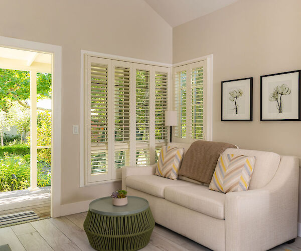 A cozy room with a white sofa, green table, wall art, large windows with blinds, and an open door to a sunny garden view.