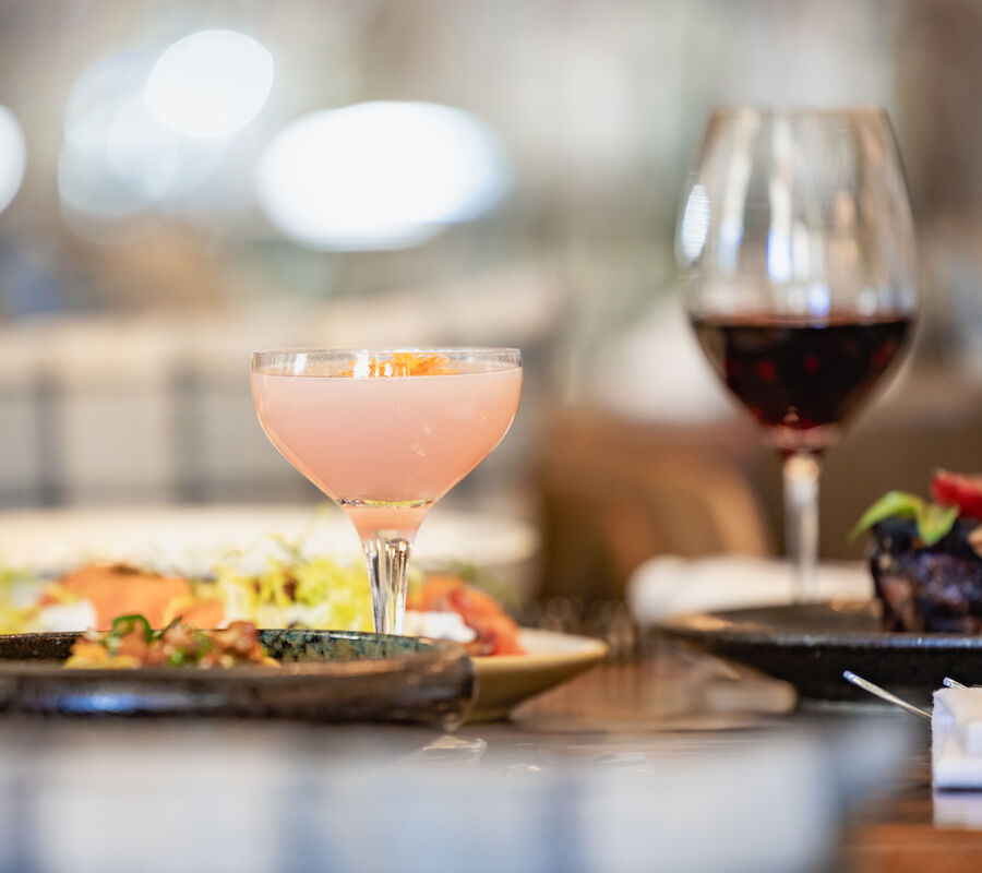 A table with a pink cocktail, a glass of red wine, and plates of food, including salad and steak, is set for dining.