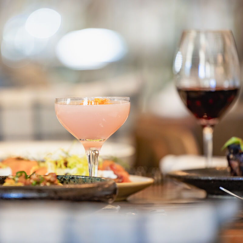 This image shows a dining table with a cocktail, a glass of red wine, and plates of food, creating a cozy dining atmosphere.
