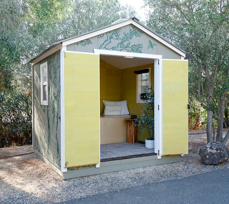 A small shed with open yellow doors, situated outdoors. Inside, there's a cozy bench with cushions and a small stool.