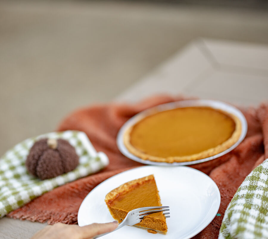 A slice of pumpkin pie on a plate is being reached for, with a whole pie and decorative elements in the background.
