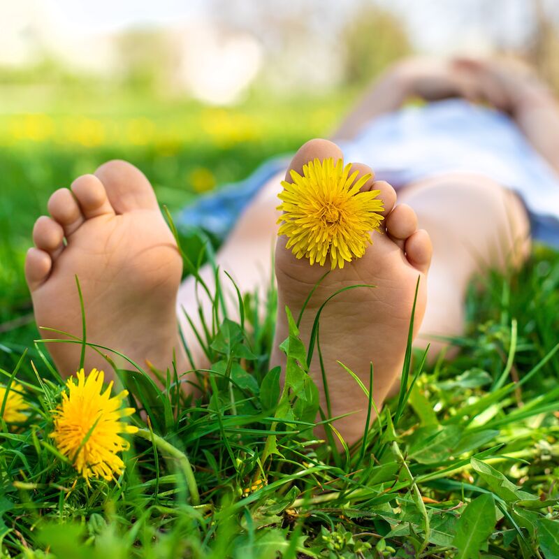 A person lying in grass with bare feet, one adorned with a yellow flower, surrounded by green grass and more yellow flowers.