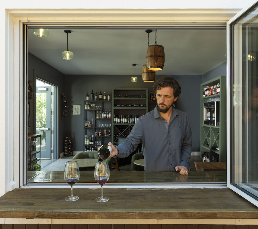 A person pouring wine in a modern room, viewed through a window, with bottles on shelves and two wine glasses on the ledge.