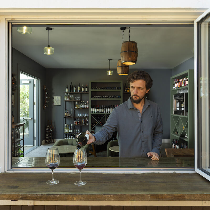 A person pouring wine into glasses at a window counter, with a view of a wine shop interior in the background.