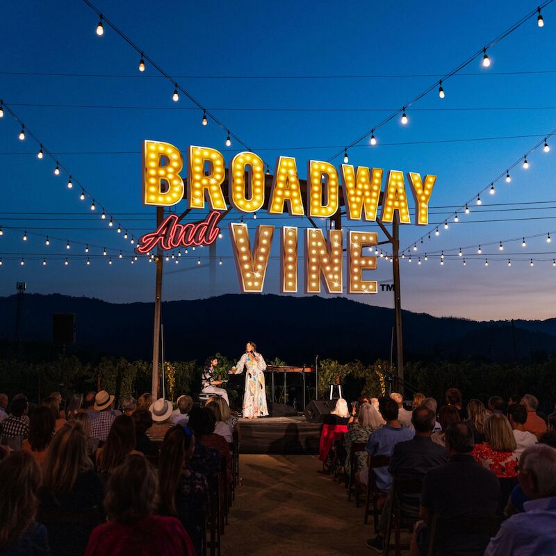 An outdoor event with a stage, audience, and marquee lights reading 