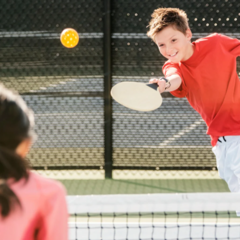 A child in a red shirt plays pickleball, hitting the ball toward an opponent on a court with a net and fencing in the background.