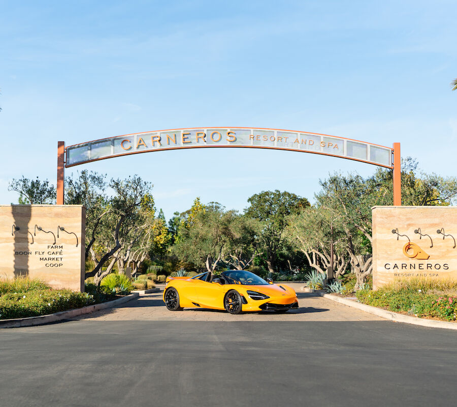 A yellow sports car is parked at the entrance of Carneros Resort and Spa, surrounded by greenery and palm trees.