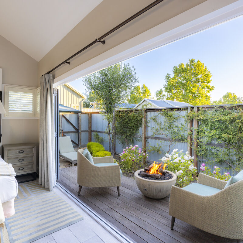 A cozy bedroom opens to a patio with two chairs, a fire pit, and lush garden views.