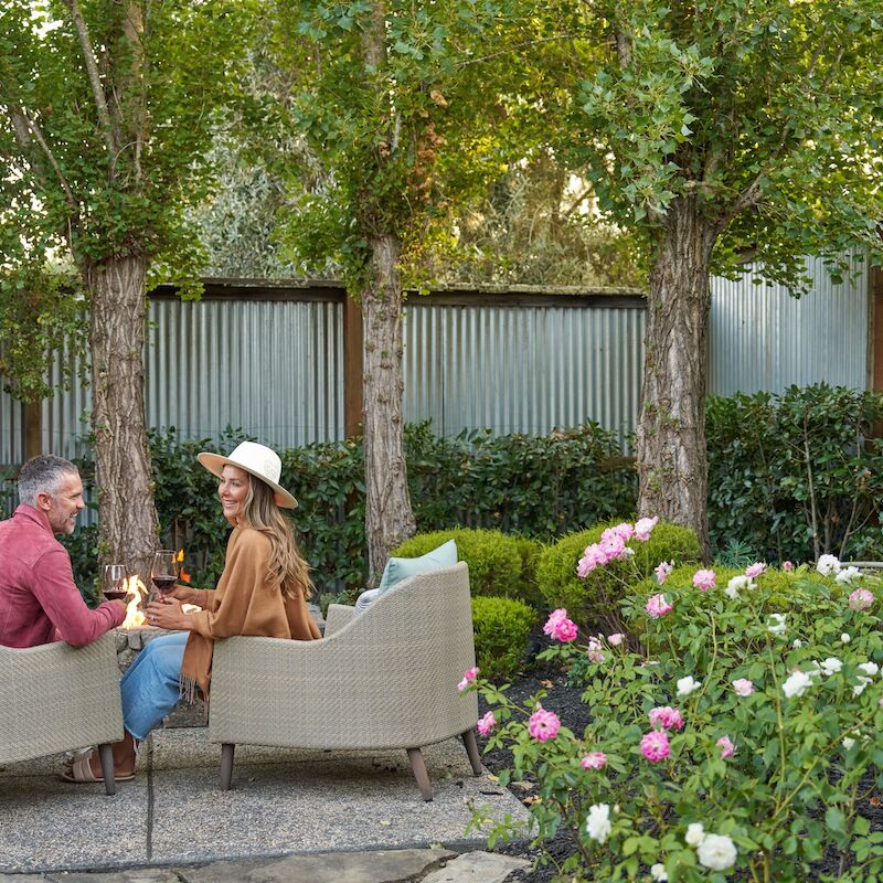 A couple sits outdoors on patio furniture, surrounded by lush greenery and colorful flowers, enjoying a peaceful garden setting.