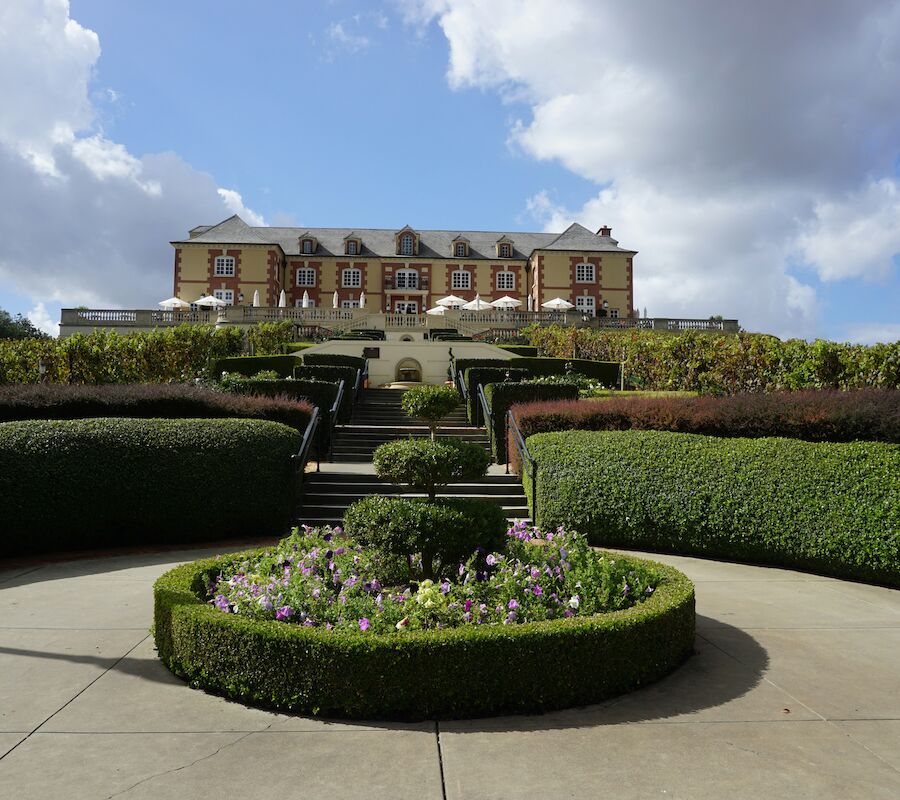 The image shows a grand building atop a landscaped hill, with symmetrical gardens and pathways leading up to it under a partly cloudy sky.