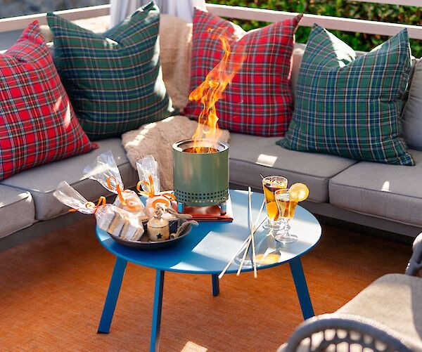 A cozy patio setup with plaid pillows, a small fire pit, treats, and drinks on a blue table surrounded by seating.