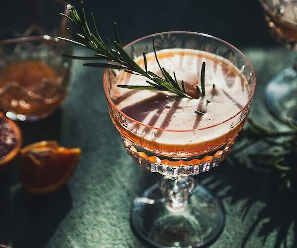 A cocktail in a crystal glass garnished with a sprig of rosemary, set on a dark surface with citrus slices nearby.