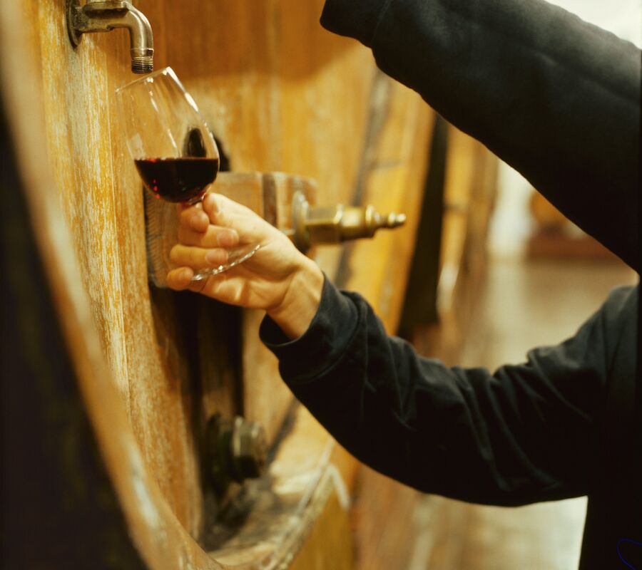 A person fills a wine glass from a wooden barrel tap in a cellar.