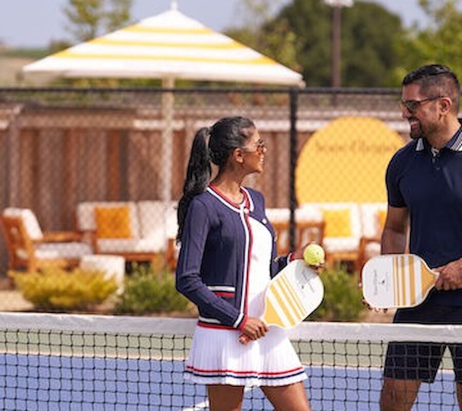 Two people are on a pickleball court holding paddles, with seating and an umbrella in the background.