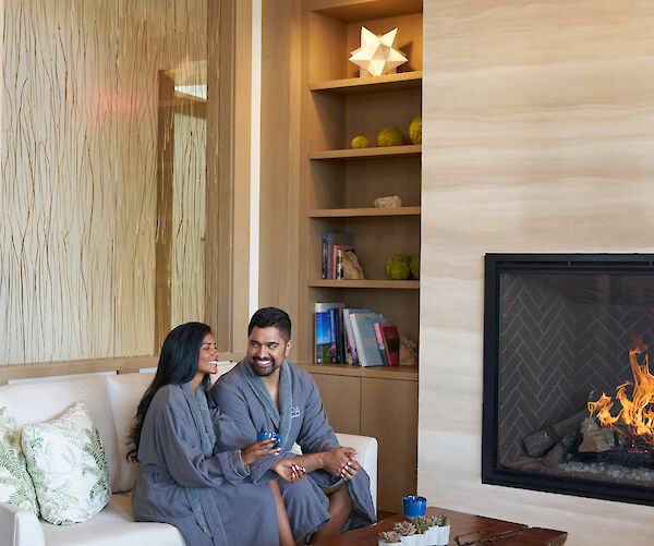 A couple sitting on a couch by a fireplace, wearing robes, in a cozy room with bookshelves and a wooden coffee table.