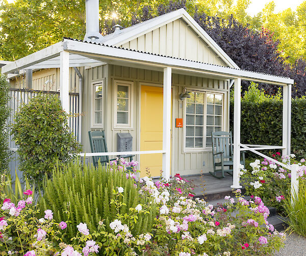 A quaint cottage with a yellow door, surrounded by colorful flowers and greenery, is seen in the image.
