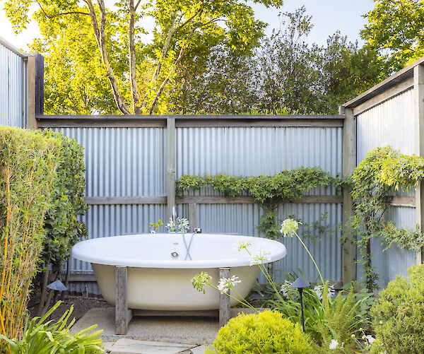 The image shows an outdoor bathtub surrounded by greenery and stone tiles, with a metal fence and trees in the background.