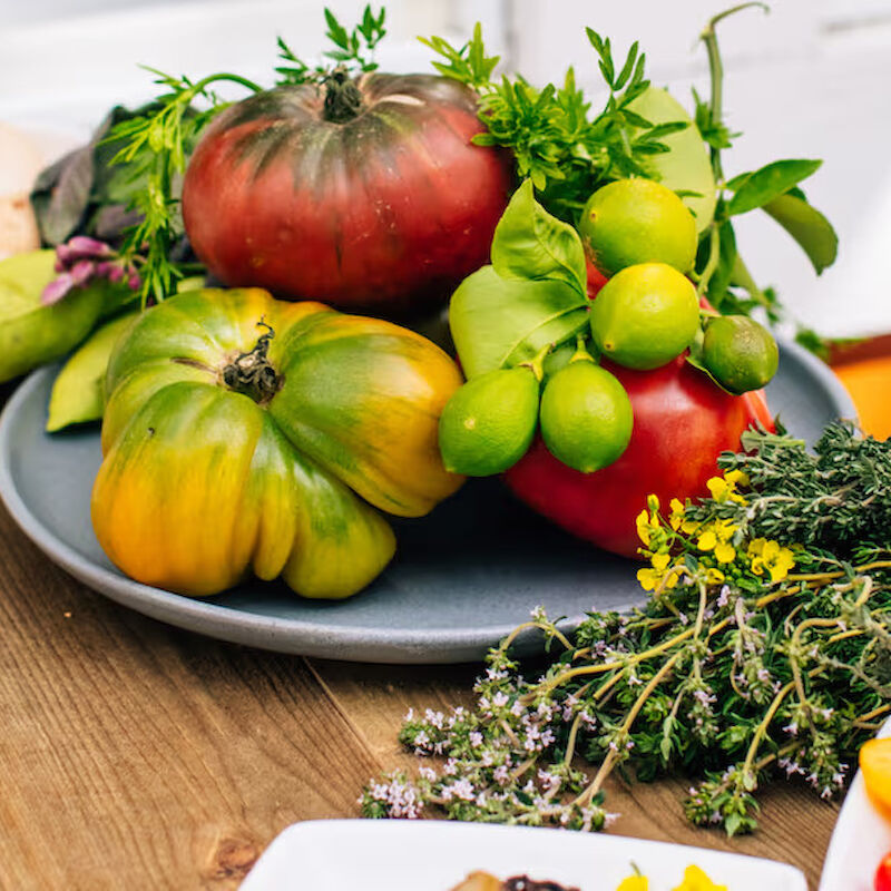 The image shows a variety of fresh produce, including heirloom tomatoes, mixed herbs, and eggs, all artfully arranged on a wooden surface.