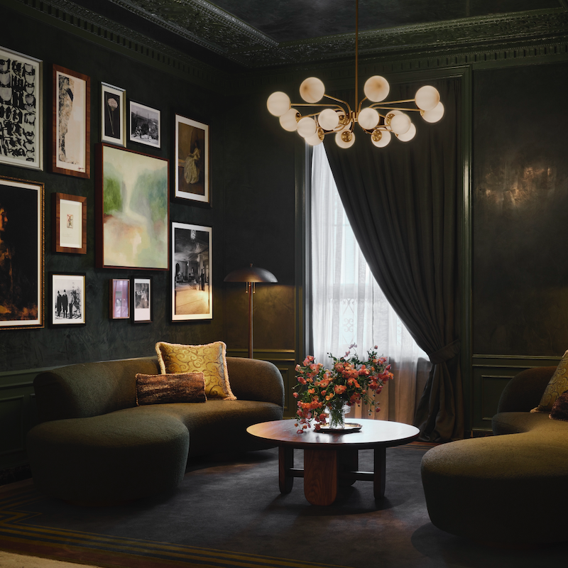 A stylish living room with dark walls, cozy seating, a round table, floral centerpiece, and a modern chandelier. Art decorates the walls.