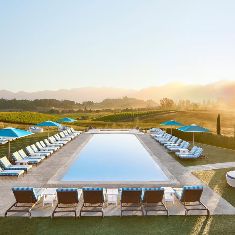 A scenic outdoor pool with loungers and umbrellas, set against a backdrop of vineyards and mountains at sunrise or sunset.