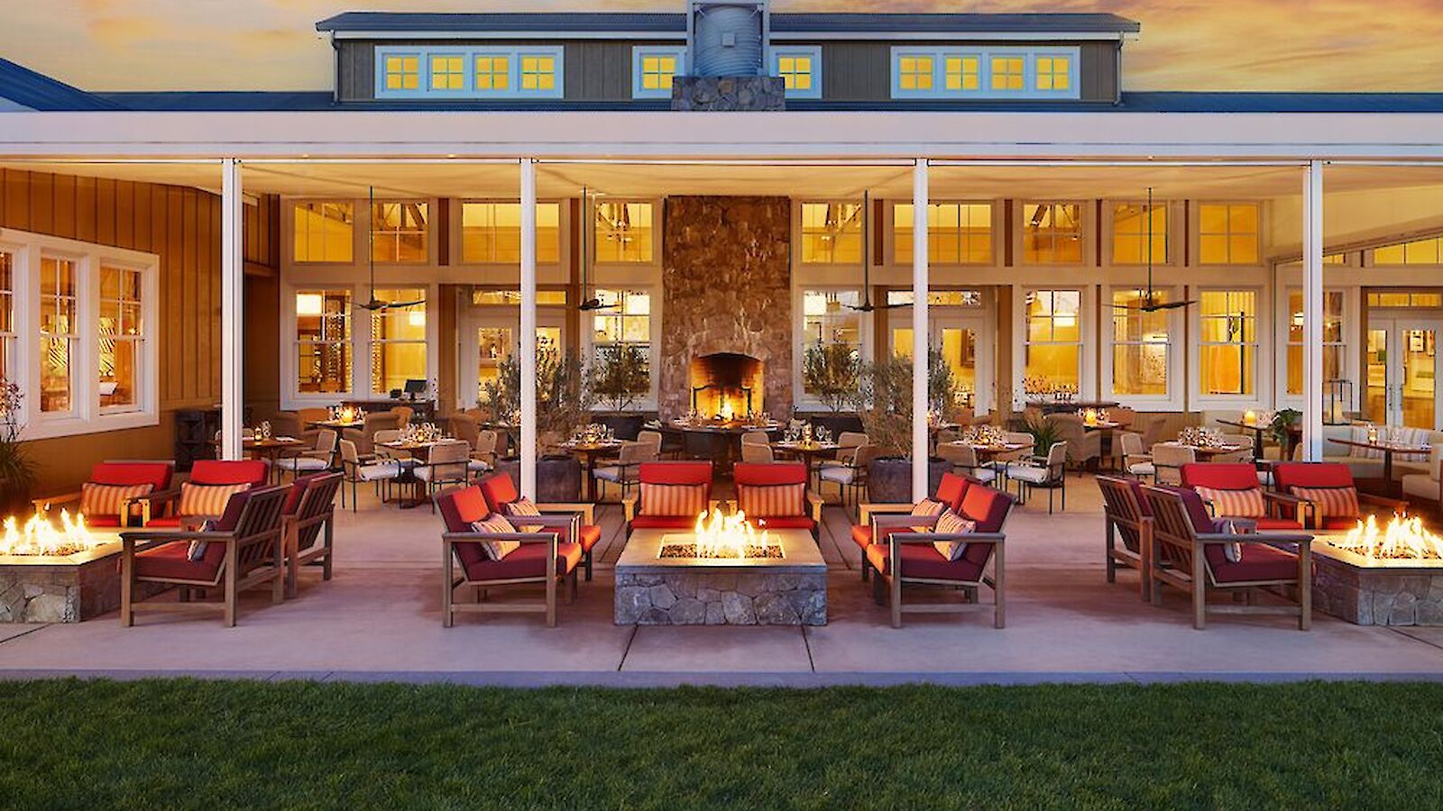 The image shows a cozy outdoor seating area with several fire pits, surrounded by chairs, in front of a building at sunset.