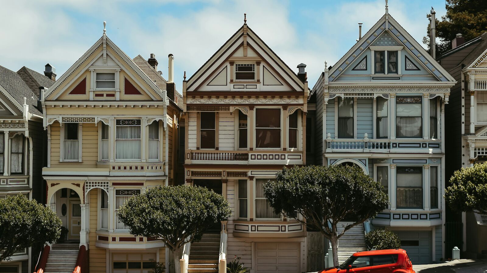 The image shows colorful Victorian-style houses with intricate details, trees in front, and a small red car parked on the street.