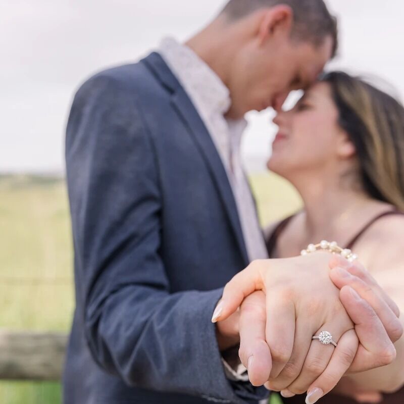 A couple embraces closely outdoors, with the focus on their hands showing an engagement ring.