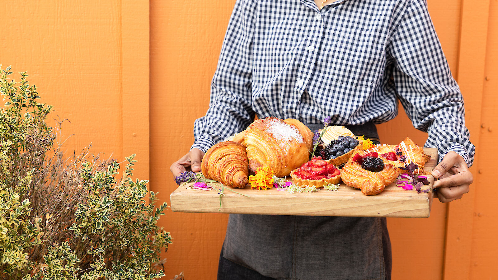 A person holds a wooden tray with various pastries and fruits against an orange background.