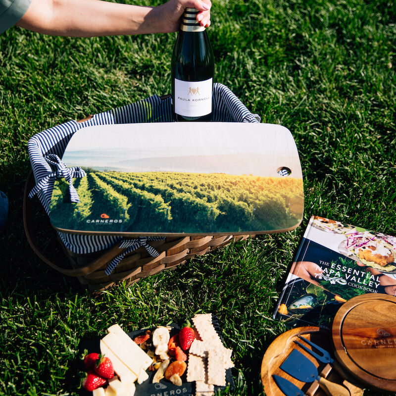 A picnic setup with wine, a basket, a magazine, and a wooden serving dish on grass.