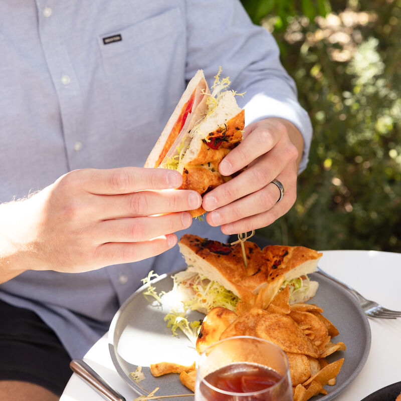 A person in a blue shirt is holding a sandwich over a plate with fries, seated outdoors with greenery in the background.
