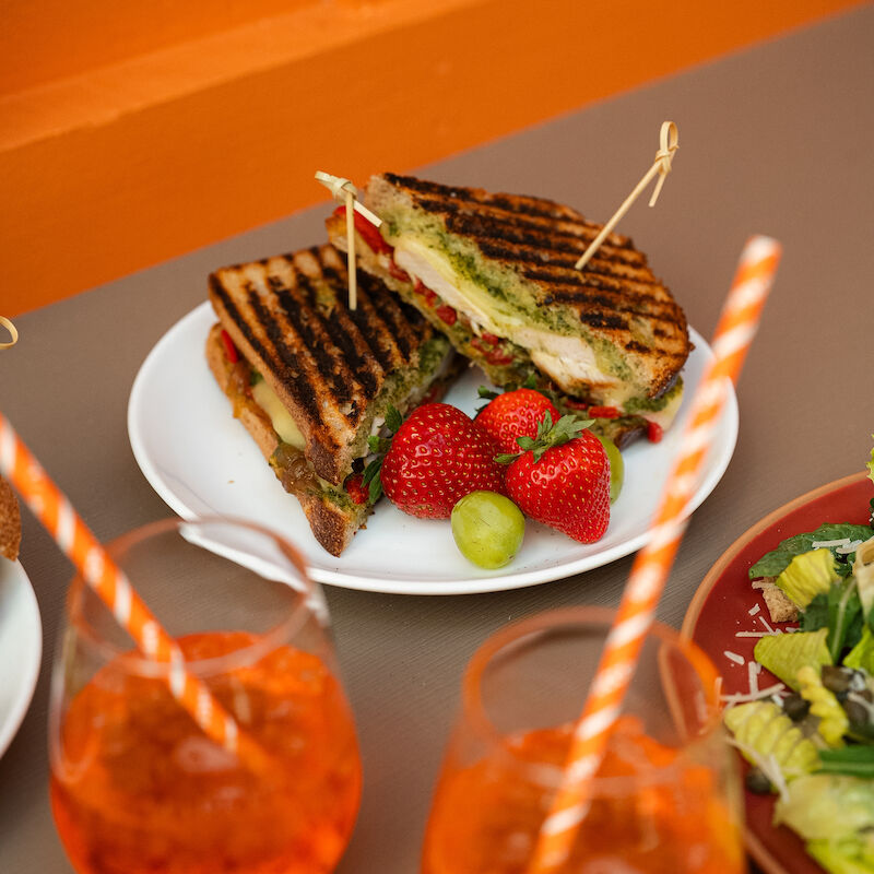 Grilled sandwiches with strawberries and grapes on a plate, accompanied by two orange drinks with striped straws, set on a table.