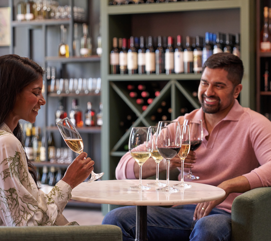 Two people enjoying a wine tasting at a table, surrounded by shelves of wine bottles, smiling and engaged in conversation.