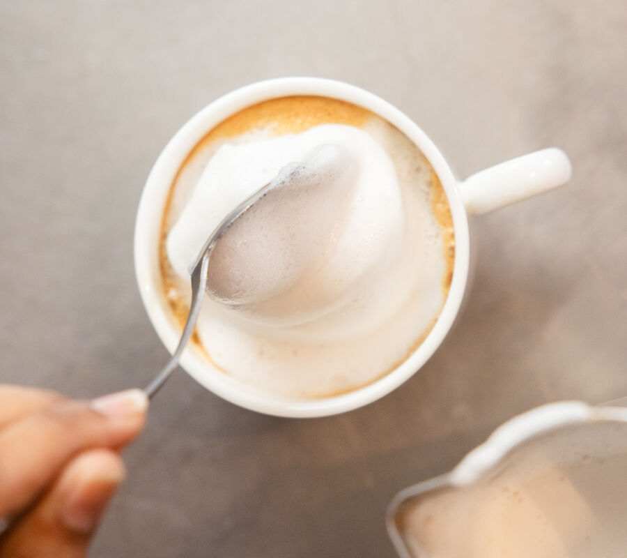 A person is using a spoon to scoop foam from a cup of coffee or cappuccino, with a milk frothing pitcher nearby.
