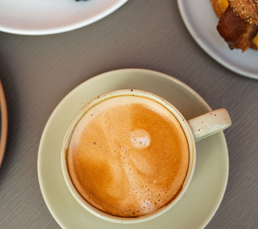 A cup of coffee with froth is in the center, surrounded by a plate of berries and flowers, and a bagel sandwich on separate plates.