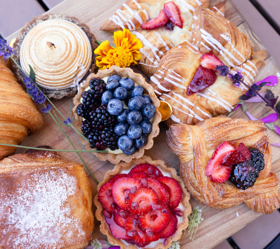 An assortment of pastries, including fruit tarts and croissants, arranged on a wooden board with edible flowers and berries.