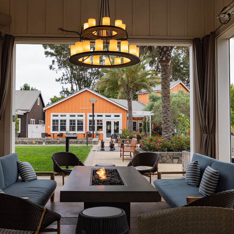 A cozy outdoor patio with a fire table, surrounded by chairs, looks out to a garden and orange building under a chandelier.