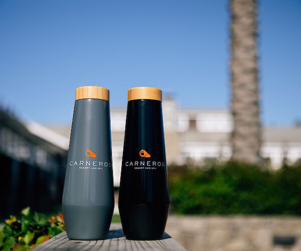 Two branded bottles with wooden caps are set against an outdoor backdrop with greenery and a building under a clear blue sky.
