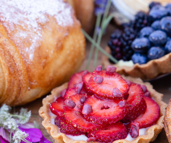 A variety of pastries, including a croissant, a tart with sliced strawberries, and a tart with blueberries and blackberries, garnished with flowers.