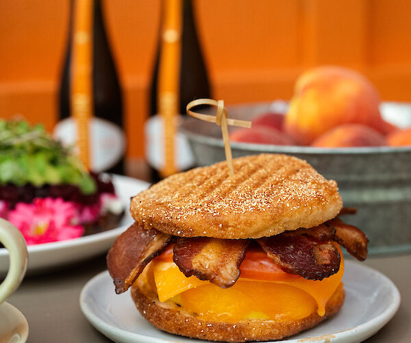 A sandwich with bacon, cheddar cheese, and tomato on a plate. There's a skewer on top and drinks, salad, and fruit in the background.