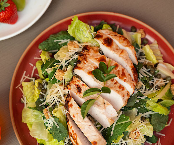 A plate of salad with sliced grilled chicken, leafy greens, and grated cheese. A side plate has strawberries and grilled bread.