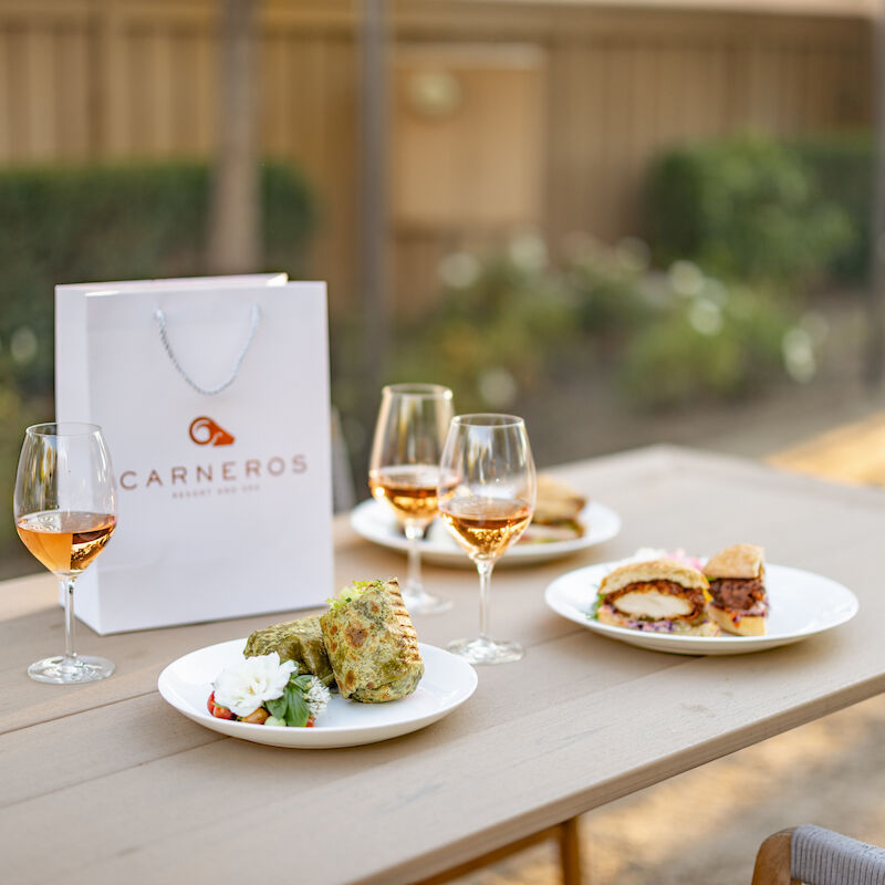 A table with plates of food, glasses of wine, and a Carneros shopping bag, set outdoors.