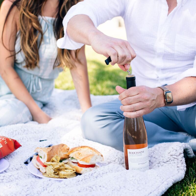 A couple is having a picnic on a blanket with chips, sandwiches, and a bottle of wine being opened.