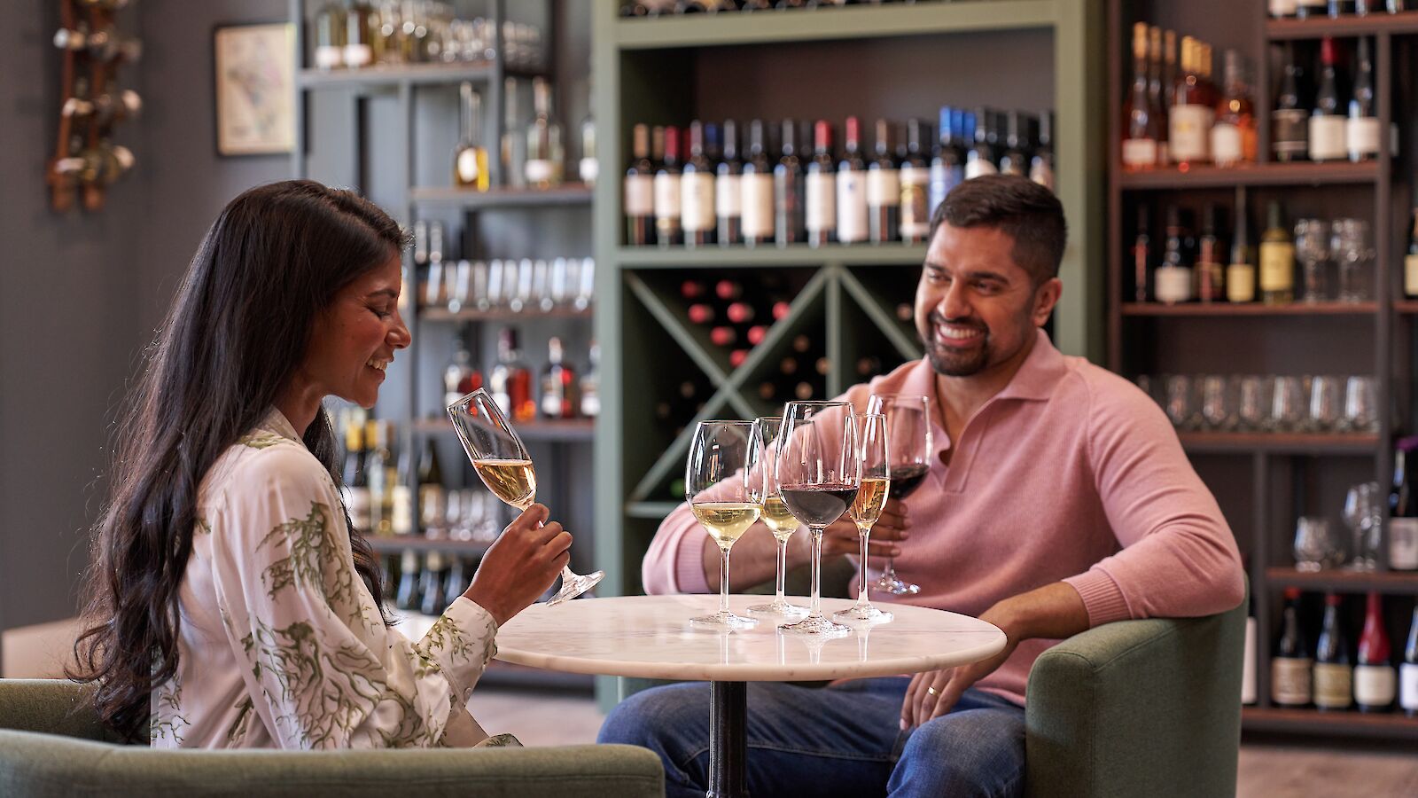 A couple enjoys wine tasting at a table in a wine shop, surrounded by shelves of wine bottles.