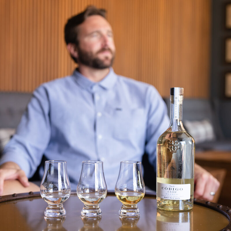 A man sits with three glasses and a bottle on a table, with a blurred background.