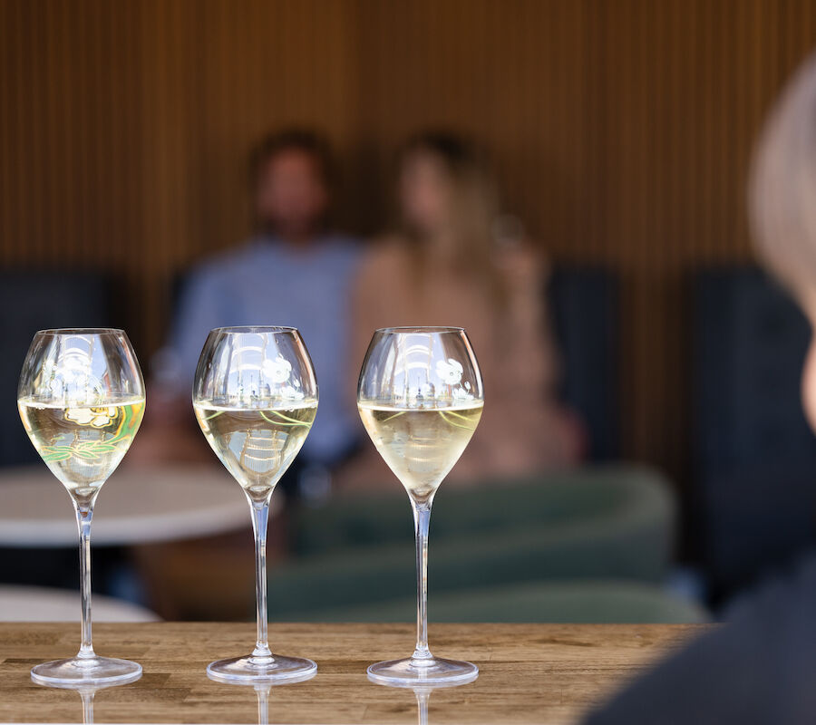 Three glasses of white wine are on a table, with people sitting in the background, slightly out of focus.
