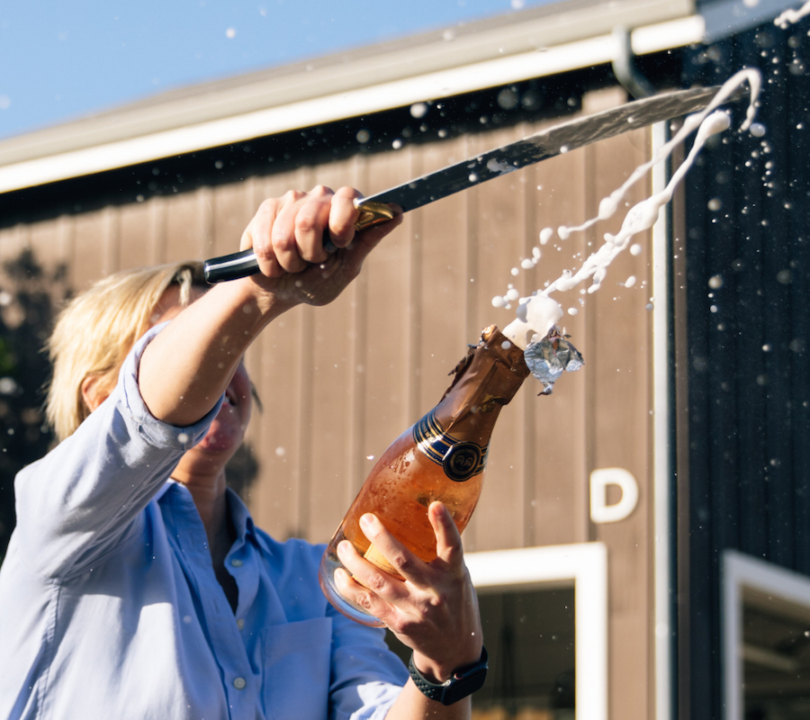 A person is sabering a champagne bottle, causing it to spray, outside a building.