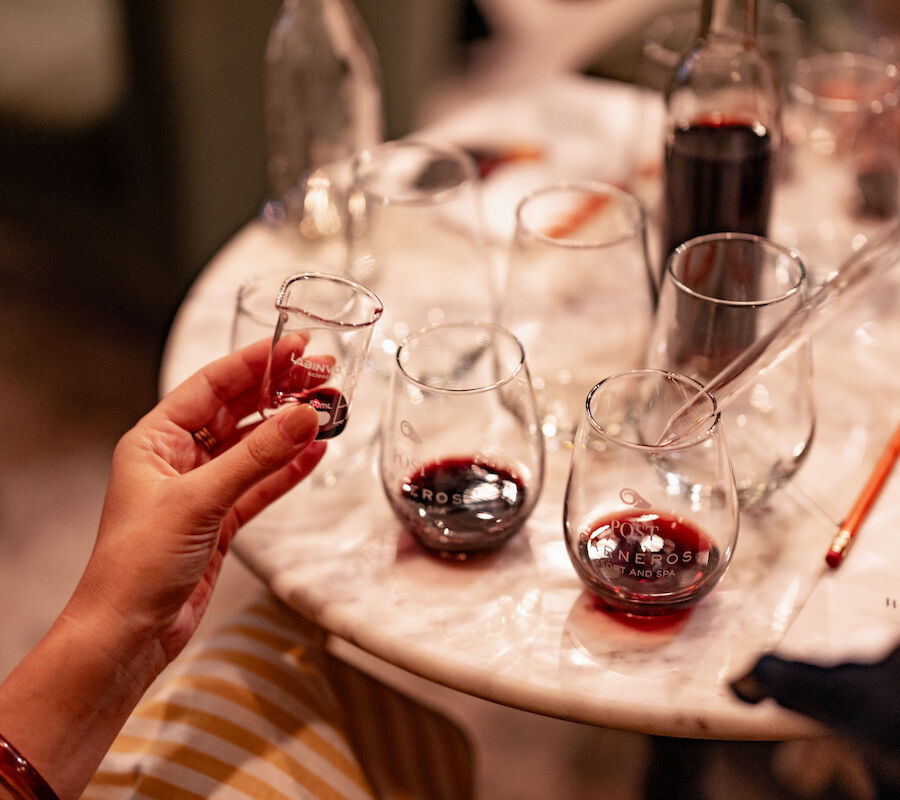 A person is holding a small glass of red liquid, with other glasses and a bottle on a round table in a cozy setting.