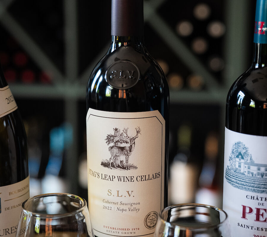 Three wine bottles and two glasses filled with red wine are displayed with a blurred background of wine bottles.