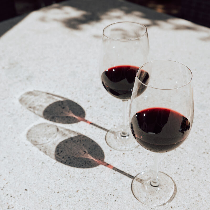 Two glasses of red wine casting shadows on a sunlit surface.