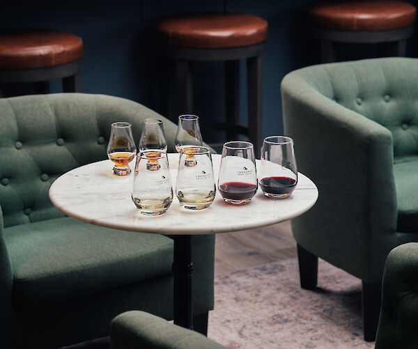 A round table with wine glasses is surrounded by green chairs in a cozy setting, featuring a bar area with stools and books.